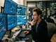A male cryptocurrency trader sits thoughtfully at a dark desk with his hand on his chin, looking at a multi-monitor setup displaying complex financial charts, real-time market data, and the MoonDog AI ($MNDG) trading platform interface in a modern city office at night.