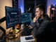 A young man sits at a desk in a dark room analyzing volatile cryptocurrency charts on dual monitors, featuring texts like "MEME COIN VOLATILITY" and "HIGH RISK," with a Dogecoin figurine on the desk.