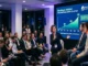 A corporate panel discussion presentation showing a MoonDog AI (MDOGAI) price prediction and growth trajectory chart on a large screen spanning from 2024 to 2030, with a speaker addressing an audience of crypto investors.