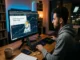 A man in a grey hoodie sits at a wooden desk at night, analyzing cryptocurrency charts and an article titled 'What Is a Meme Coin? The Complete Guide' on a large computer monitor. An illustration of a Dogecoin-style Shiba Inu in an astronaut suit overlays the financial graphs.