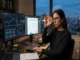 A female crypto analyst examining a physical utility token with a stylized 'U' logo at her desk, with dual monitors displaying cryptocurrency trading charts and a token utility evaluation framework against a city skyline at dusk.