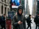 A concerned young man in a grey hoodie checks his smartphone displaying a crashing red Bitcoin chart on a busy, overcast city street.